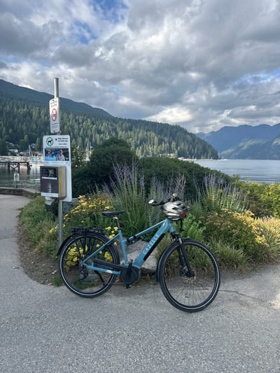 An electric bicycle, a blue Gazelle model, is parked on a paved path in front of a lush garden with yellow and purple flowers. The bicycle has a black rack and a silver helmet is placed on its handlebars. In the background, a calm body of water, likely an inlet, is dotted with sailboats. The water is bordered by a dense, tree-covered mountainside, characteristic of the North Shore mountains in North Vancouver, Canada. A signpost with various notices, including "No Smoking" and "Dogs allowed on leash only," stands to the left of the bicycle. The sky is partly cloudy, with dramatic grey clouds interspersed with patches of blue. The scene appears to be a peaceful, scenic spot, possibly a park or waterfront pathway.