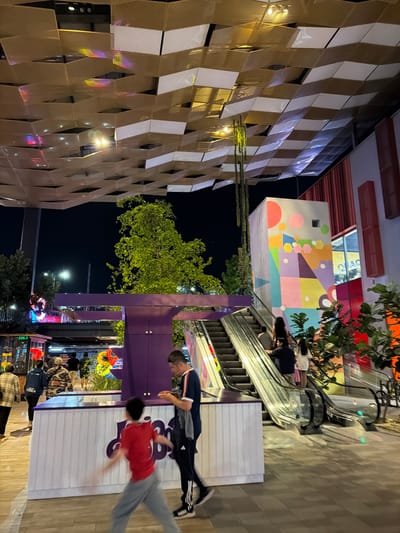 The image shows an outdoor scene in San Pedro Cholula, Mexico at night. A tall building with a modern, geometric facade of beige and white panels dominates the left side of the frame. Adjacent to it is a multi-story building with a colorful, abstract mural on one wall. An escalator leads up to the mural wall.

Two men are walking on a pathway in the foreground. One is wearing a red shirt and light gray pants, and the other is wearing a blue jacket and black Adidas pants. Several other people are milling about, some appearing to be looking at merchandise. The environment is illuminated by streetlights.