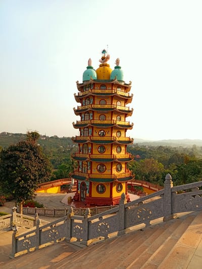 The image shows a tall, colorful pagoda with multiple tiers, likely a Buddhist temple. The structure is predominantly yellow and red, with decorative green and gold accents. Each tier features windows, and the topmost tier is crowned with yellow and green domes. The pagoda is situated on a raised platform, surrounded by trees and greenery, giving the impression that it is located on a hill or elevated terrain. In the foreground, there are stone stairs and ornate railings leading up to the pagoda, suggesting a path for visitors. The background consists of a hazy sky and distant hills or mountains. The picture appears to be taken during the day, with soft, natural lighting. Based on the information provided, it is located in the city of Phú An, Vietnam.