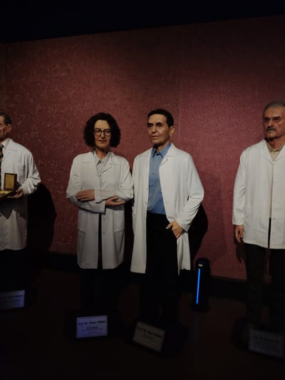 This image captures an indoor exhibit featuring four wax figures of prominent scientists, likely in a museum setting in Eskisehir, Turkey. All four figures are dressed in white lab coats and are positioned in front of a dark red wall adorned with a subtle leaf-like pattern. The lighting is low, casting shadows on the dark floor.
From left to right, the first figure is a man with glasses, holding an open box containing a gold-colored rectangular object, possibly a medal. His plaque is partially visible, reading "Prof. Dr. Aziz SANCAR".
Next is a woman with dark, curly hair and glasses, wearing a patterned shirt under her lab coat, with her arms crossed. Her plaque clearly states "Prof. Dr. Özlem TÜRECİ". Below her name, it reads "BİLİM İNSANI" (Scientist), "mRNA Aşı Teknolojisi-Biontech kurucusu" (mRNA Vaccine Technology - BioNTech founder), and "Nobel adayı" (Nobel candidate).
Adjacent to her is a man with dark hair, wearing a blue shirt under his lab coat, with one hand in his pocket. His plaque identifies him as "Prof. Dr. Uğur ŞAHİN", also noted as a "BİLİM İNSANI" (Scientist) and "mRNA Aşı Teknolojisi-Biontech kurucusu" (mRNA Vaccine Technology - BioNTech founder). A blue glowing cylindrical light is visible on the floor between this figure and the woman.
The final figure on the right is an older man with a mustache and glasses, wearing a light-colored shirt under his lab coat. His partially visible plaque appears to read "Prof. Dr. Naci ELİK". The figures are posed statically, conveying a sense of honor and recognition for their scientific achievements.