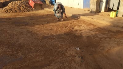 The image shows a woman in a floral dress sweeping a dirt road in Kayna, Democratic Republic of Congo. The road is covered in dirt and has a few puddles of water. There is a pile of dirt and a red container on the left side of the image and a wooden structure and two yellow containers on the right side. There is a blue wall behind the wooden structure.  The woman is sweeping towards the right of the image with a broom. The ground is dusty and uneven, with a few small plants growing out of the ground. The scene is a typical street scene in a developing country.