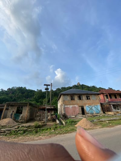 This image captures a street scene in Igarra, Nigeria, dominated by natural landscape and human structures under a partly cloudy sky. The foreground, at the bottom right, is partially obscured by a human finger. A paved road curves through the lower portion of the frame.

On the left side, a partially constructed or dilapidated concrete block building features exposed blockwork and an open window-like aperture. A small pile of sand rests in front of this structure. A male individual, wearing a striped shirt and dark trousers, stands near the sand pile, facing away from the camera.

In the center, a tall utility pole, possibly made of wood or concrete, supports a solar panel assembly near its top. Wires extend from this pole and across the scene horizontally. Beyond this pole, more individuals are visible, including one seated person and another standing further back.

On the right side of the frame, a two-story building with a dark-colored hip roof is prominent. Its facade shows significant weathering with peeling paint in hues of yellow, pink, and light blue. Several windows are boarded up or open, and blue wooden shutters or boards cover sections of the lower level. Adjacent to it, a second two-story building with a reddish-brown roof and facade is partially visible, also appearing worn.

The background is characterized by lush, green, heavily forested hills or mountains. Distinctive rock formations are visible on the summits of these hills, particularly a prominent, somewhat conical rock on the rightmost hill and another larger rock on the left. The sky above is light blue with scattered white and grey cumulus clouds. Power lines stretch across the upper mid-ground. The ground between the road and buildings is a mix of dirt and sparse vegetation, with additional small piles of sand visible.