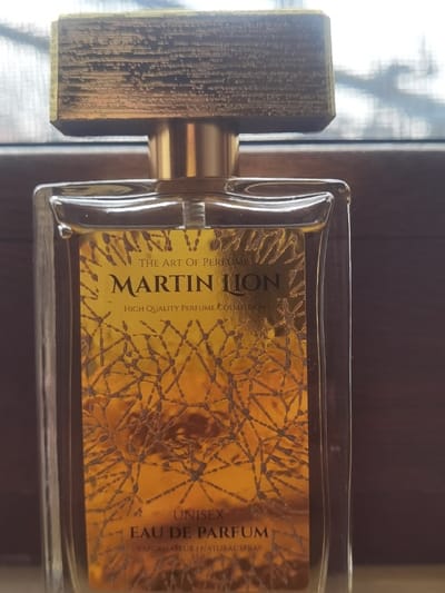 A close-up, vertically oriented photograph displays a clear glass perfume bottle positioned centrally. The bottle is rectangular with subtly rounded edges and contains a transparent liquid. It features a golden-colored atomizer component and is capped by a rectangular, gold-toned cap with a distinct, rough, wood-grain texture. The front of the bottle is adorned with a golden-yellow label featuring a dark, intricate, fractal-like pattern. Text on the label reads "THE ART OF PERFUME" at the top, "MARTIN LION" prominently below, followed by "HIGH QUALITY PERFUME COLLECTION," and near the bottom, "UNISEX," "EAU DE PARFUM," and "VAPORISATEUR | NATURAL SPRAY." The background is out-of-focus, showing a dark horizontal element, likely a window frame, behind the bottle, and a brighter, diffused light source originating from a window, with faint, blurry dark shapes suggesting outdoor foliage. The scene is located in the city of Velichkovo, Bulgaria. No individuals or actions are depicted.