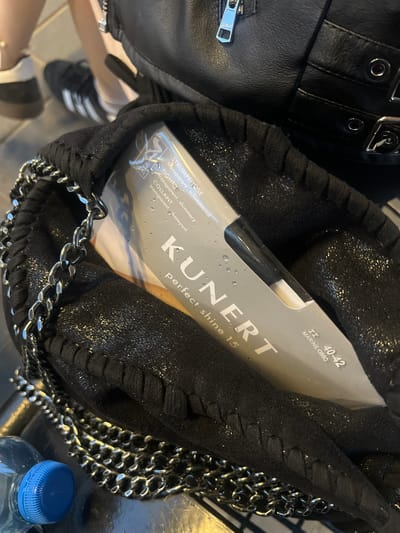 The image shows a partially visible package of KUNERT brand tights inside a black, textured handbag with a chain strap. The packaging indicates the tights are "perfect shine 15", transparent, size II (40-42), and the color MARINE/0880. The handbag's lining appears to have a sparkling effect. Part of a black leather garment with metal buckles and a zipper is visible to the left. The background includes a glimpse of a person's legs, wearing white socks and black Adidas sneakers. A plastic water bottle with a blue cap is also in the frame. The overall composition suggests the photo was taken indoors, possibly in Beverwijk, Netherlands, although specific location details are not evident.