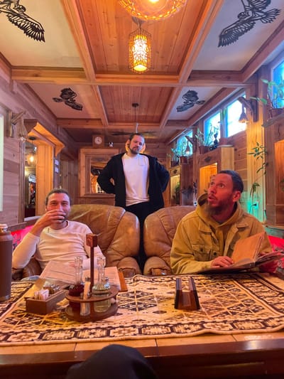 The image, correctly oriented, displays three men relaxing in a warm, rustic indoor setting, likely a cafe or lounge, in Moračko Trebaljevo, Montenegro. The room is extensively paneled with warm-toned wood on the walls and ceiling, featuring visible wooden beams.

Natural light enters through skylights set into the ceiling, where green foliage of plants is visible. This is complemented by the warm glow from several decorative light fixtures, including a large, round woven pendant and a hexagonal metal lantern. The walls are adorned with framed white panels showcasing black metal silhouettes of birds, possibly eagles or hawks, in flight. A patterned rug with geometric designs covers the floor area, adding to the cozy atmosphere. A wall clock is also visible in the background.

Three men are present. In the foreground, a man with a beard, wearing a white long-sleeved shirt, is seated, leaning forward with his hand near his mouth, looking towards the camera with a thoughtful expression. To his right, another man, also bearded, in a tan jacket, is seated on a plush light brown leather armchair. He holds a menu or book and looks at the camera with an open-mouthed, expressive, perhaps surprised or playful gaze. Further back, a third man in a white t-shirt and black jacket stands or leans, looking directly at the camera.

On the wooden table in front of the seated men, a circular wooden caddy holds several glass bottles, likely for beverages or condiments, alongside a small wooden box. The overall scene conveys a relaxed, social gathering in a comfortably furnished space. The time of day appears to be during daylight hours due to the skylights, enhanced by the warm artificial lighting. No discernible text is visible besides the potential menu.