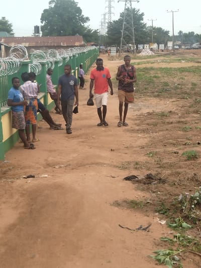 The image shows a scene in Kanshio, Nigeria, at the coordinates 7.683° N, 8.537° E. There are seven people visible. Five of them are standing. One is a young boy, wearing a blue shirt and carrying a bag on his back. He is standing next to a young girl, wearing a blue dress. Another young girl, wearing a striped dress, is standing next to her. There is a man wearing a red shirt and khaki pants, standing next to another man, wearing a black sleeveless shirt and a black cap, and a third man is wearing a blue shirt and black pants. The scene appears to take place on a dirt road, in front of a green fence with barbed wire at the top. The fence runs along the left side of the image. The road is surrounded by brown dirt and sparsely scattered green vegetation. In the background, there are power lines, trees, and a few cars. The ground is mostly dry and dusty, with patches of green vegetation. The area looks to be outside and near a residential or commercial area.