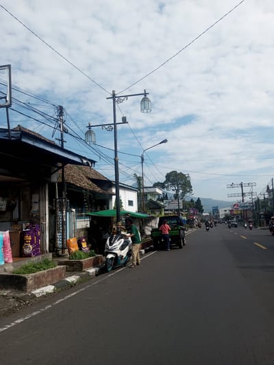 The image depicts a street scene in a Southeast Asian city, possibly Zeyathiri District, Myanmar. The road is paved and appears to be two lanes. On the left side of the road, there's a building with a decaying roof and a storefront displaying bags of animal feed and other items. Adjacent to the store, there's a person standing beside a white scooter with blue accents. Nearby, a small green pick-up truck is parked with a woman standing near the back. The right side of the road shows moderate traffic consisting of motorcycles and cars. Further down the road, there are streetlights and commercial signs. The sky is partly cloudy. Mountains are visible in the distance. Overhead power lines run across the street.