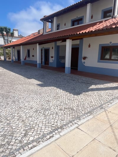 A wide-angle, ground-level outdoor shot captures the facade of a two-story building with a covered walkway. The building features white walls with a light blue trim accenting the windows and roofline. Red clay tiles cover the roof, and brown wooden beams support the overhang of the walkway. The walkway itself is paved with small, irregular grey and white stones, casting distinct shadows from the building's architecture. To the right, the paved area transitions to larger, light-colored square tiles. A row of palm trees is visible in the background to the left, and a clear blue sky with scattered white clouds forms the backdrop. The scene appears to be in a residential or commercial area, possibly a hotel or guesthouse, located in Caldas da Rainha, Portugal. There are no people or animals present in the image.