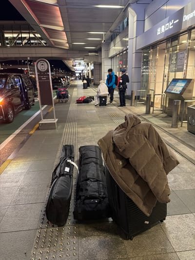 The image captures a luggage-laden scene at an airport or transportation hub. A large suitcase with a brown corduroy jacket rests on top, followed by two black duffel bags, possibly for skis or snowboards. People stand in the distance near a brightly lit departure area. Taxis line up outside, with a prominent sign indicating transportation options. The overall scene suggests travel preparations and departure.