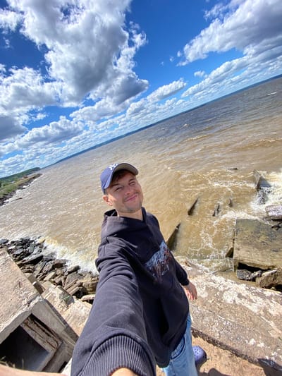 The image depicts a young man taking a selfie by a body of water. He is wearing a dark hoodie, blue jeans, a blue baseball cap, and purple Crocs. The man is smiling, and the scene is brightly lit, suggesting it is daytime. The water appears murky and choppy, with visible waves and some debris along the shore, which is made of concrete and rocks.
The background is dominated by a bright blue sky filled with fluffy white clouds. The perspective of the photo is tilted, with the sky taking up a significant portion of the frame on the left and the water on the right. This unusual angle makes the horizon appear diagonal.
There is no visible text in the image. The overall impression is of a casual outdoor moment, possibly at a waterfront park or industrial area, given the concrete structures. The weather appears sunny.