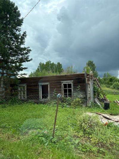 The image depicts an abandoned wooden house located in Крынкаўскі сельскі Савет, Belarus. The house is in a state of disrepair, with a partially collapsed wall on the right side. Several windows are broken or missing frames. Overgrown vegetation surrounds the structure, with tall grass and weeds covering the ground. A fence runs in front of the house. In the background, a dense line of trees forms the horizon, and the sky is filled with dark, heavy clouds, suggesting an impending storm. A lone power line stretches across the upper portion of the image. There are no people or animals visible in the scene. The environment appears rural and neglected.