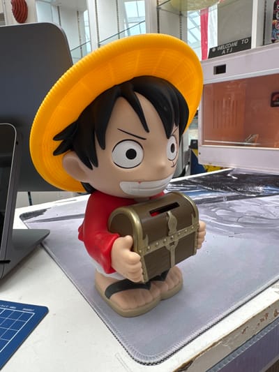 A close-up, eye-level, medium shot captures a cartoon-like piggy bank shaped like the character Monkey D. Luffy from the anime One Piece. The piggy bank is predominantly yellow for the hat, red for the shirt, and beige for the skin and feet, wearing black sandals. It holds a small, brown treasure chest with gold accents, which serves as the coin slot. The character has large, round eyes with black pupils and an open-mouthed smile.
The piggy bank is placed on a light gray mousepad with a textured surface. Behind the character, a large, dark gray, angled object, possibly a monitor or laptop screen, is visible on the left. In the background, a white counter or table supports various items, including a white and beige box with a glass front, inside which a small reptile, likely a lizard or gecko, is perched. A black sign above this box reads "WELCOME TO ATJ". The overall environment suggests an indoor retail or display space, potentially a gift shop or electronics store. The lighting is bright and natural, coming from above, indicating a well-lit interior, possibly with large windows or a skylight. The image composition focuses on the piggy bank as the central subject, with the background providing context for its display. There is no indication of the specific location within El Paso, United States of America.