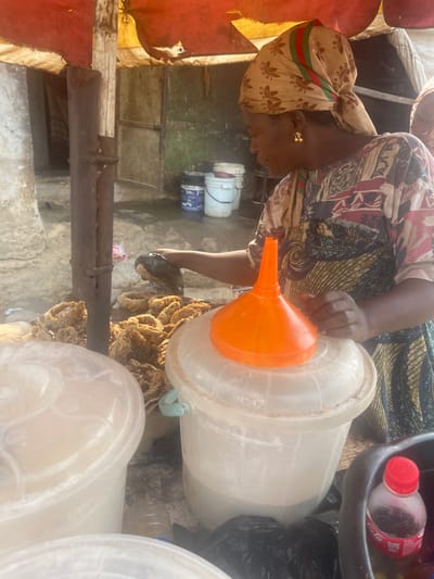 The image depicts an outdoor food stall in Dzhidu, Nigeria. A woman, estimated to be middle-aged, is positioned slightly right of the frame's center, engaged in an activity involving food preparation or handling. She wears a multi-colored patterned dress and a beige headscarf adorned with a green, red, and beige stripe pattern and Adidas logos. Gold-colored earrings are visible on her left ear. Her hands are in the process of handling a dark, irregular object, possibly food.
To her left, a quantity of light brown, ring-shaped fried food items are piled on a surface. In the immediate foreground, two translucent plastic buckets with lids are visible. An orange plastic funnel rests upside down on the lid of the bucket to the right. A small red-capped bottle, possibly containing a beverage, is visible in a dark container to the lower right.
The environment features a rustic, temporary structure with a red and orange fabric canopy supported by wooden poles. The background shows a light-colored wall with some visible wear, and several other plastic buckets, some blue, are present on a slightly elevated surface or ledge. The lighting suggests daylight conditions.