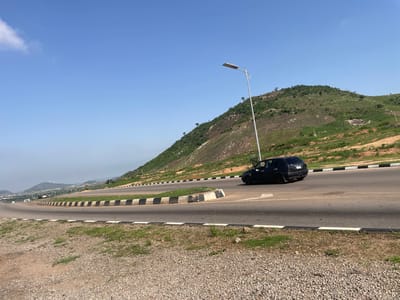 The media file captures a scenic view of a winding road in Angwan Sayawa, Nigeria. A black car is seen driving on the right side of the road, heading towards the right of the frame. The road is flanked by a concrete barrier with black and white stripes.

In the background, a lush green hill rises, dotted with trees and rocks. The sky is a clear, bright blue with a few wispy clouds. A modern streetlamp stands tall on the left side of the road, illuminating the scene. The time of day appears to be midday, with the sun casting shadows on the road and the hill.

The overall impression is one of a peaceful, natural landscape with man-made infrastructure seamlessly integrated. The location cues, such as the type of vegetation and the road design, are consistent with a tropical or sub-tropical region. There is no visible text in the media file.