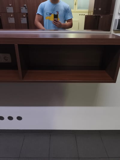A person is reflected in a mirror, wearing a light blue t-shirt with a yellow cartoon duck design and holding a yellow smartphone. They are standing in front of wooden lockers, some of which are open revealing their interiors. Below the mirror is a dark brown wooden shelf with a visible electrical outlet on the left side. The wall behind the shelf has three circular vents. The floor is tiled with dark gray square tiles. The image appears to be taken in a locker room, possibly in a public facility like a swimming pool or gym. No specific location in Pegnitz, Germany can be identified from the image.