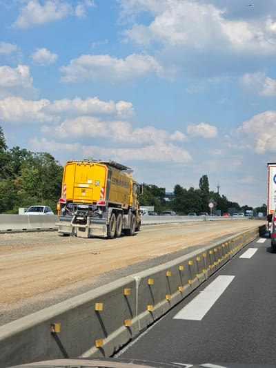 The image shows a yellow construction vehicle with "LEONHARD WEISS" written on its side, operating on a highway under construction in Rüsselsheim am Main, Germany. The highway is undergoing road work, with a new lane being constructed. The vehicle appears to be a water truck or a similar piece of heavy machinery used for road construction. In the background, there are trees and a cloudy sky. The overall scene depicts an active construction site on a highway.