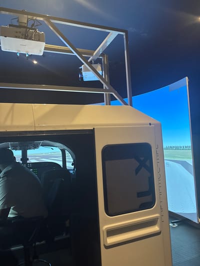 The scene captures the interior of a flight simulator facility located in Port Alfred, South Africa. In the immediate foreground to the right, the white external fuselage of a flight simulator cockpit is visible, featuring a dark rectangular window with vertical lettering, possibly "XLT," and a horizontal recessed handle below it. To the left, an open section of the simulator reveals its interior. A male individual, viewed from behind, is seated in the left pilot's seat, wearing a light-colored, short-sleeved collared shirt. The simulator's internal screens display an airport runway environment with visible markings and controls.

Above the simulator, a light-colored metal truss framework is suspended, supporting two white ceiling-mounted projectors. The projector on the left clearly displays the "Epson" brand name. The projector on the right is also white. These projectors cast a wide, panoramic image onto a large, curved projection screen positioned to the right of the simulator. The screen displays a simulated outdoor environment featuring an airport runway, green fields, a distant line of trees, and a clear blue sky. The room is generally dimly lit, enhancing the immersion of the simulated environment.