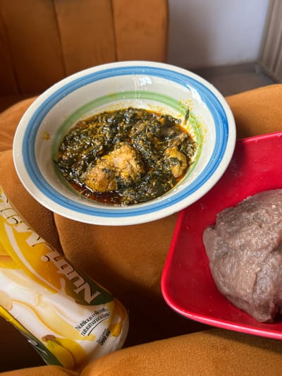 A meal is presented indoors in Egbe, Nigeria. The scene features a white ceramic bowl with blue and green striped rim, containing a dark green leafy soup or stew with several pieces of cooked poultry and reddish oil. To the right, a rectangular red plastic plate holds a single lump of brownish-purple swallow food, consistent with amala or fufu. A 500ml "NutriMilk" banana-flavored milk drink in a yellow and white flexible package is positioned to the lower left, displaying text "Nutritious milk with natural banana taste!". All items rest on a textured, orange-brown upholstered surface, likely a sofa or chair, with a similar upholstered surface and a light-colored wall visible in the background. No individuals are present.