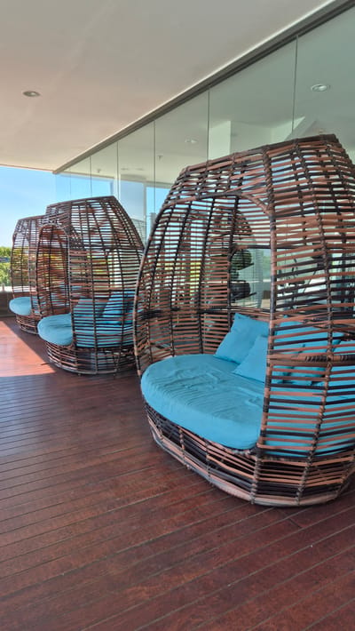 The image shows three large, spherical wicker chairs with blue cushions, arranged in a row on a wooden deck. The chairs are open, inviting relaxation. The background includes a building with a white wall and glass panels, reflecting the sky. The scene appears to be an outdoor lounge area, possibly at a resort or hotel. Given the prompt, this may be located in Banyuwangi, Indonesia, although this cannot be confirmed from the image alone. There are no people visible. The image is taken at a low angle, looking upwards towards the chairs and the building.