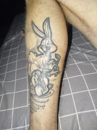 A person's lower leg features a black ink tattoo depicting Bugs Bunny, Daffy Duck, and a Tasmanian Devil-like character. The tattoo is positioned on the outer side of the leg, with the characters appearing to be in a swirling vortex. The background is a grey and white checkered bedsheet. The image suggests an indoor setting. Location information is absent.