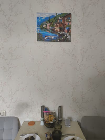 A painting of a waterfront village with boats is displayed on a wall with floral wallpaper. Below the painting, a wooden table is set for a meal, with plates, cutlery, and food items visible. Two chairs are placed at the table, suggesting a dining area. A light switch is visible on the left wall. The overall scene appears to be an indoor domestic setting.