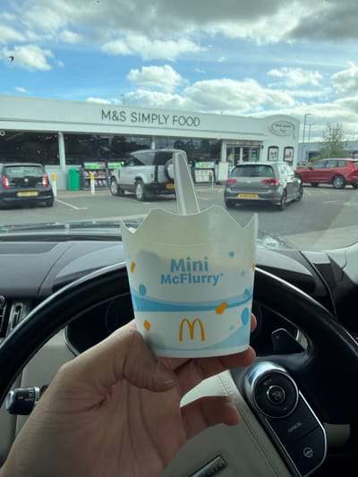 A person is holding a Mini McFlurry cup from McDonald's inside a car. The car is parked in front of an M&S Simply Food store, identified by the signage on the building. The background shows several cars parked in a lot, and a red car is visible on the right. The sky is cloudy with patches of blue. The M&S Simply Food is located at a petrol station, as indicated by the presence of fuel pumps and a "Wild Bean Cafe" sign. The scene appears to be a typical roadside convenience store and cafe. The location can be identified as the M&S Simply Food at the BP petrol station on Newport Road, Caldicot, NP26 5FU, United Kingdom.