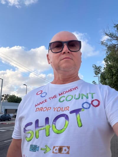 A bald man wearing sunglasses and a white t-shirt with colorful text and graphics stands outside. The t-shirt reads "MAKE THE MOMENT COUNT DROP YOUR SHOT" with stylized graphics. In the background, there are power lines, streetlights, trees, a building, and a street with parked cars, suggesting an urban or suburban environment. The sky is blue with scattered clouds. The location is identified as Even Yehuda, Israel.