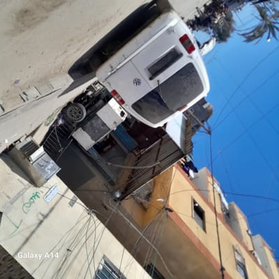 The image depicts a street scene in Khan Yunis, location unknown. A white Volkswagen van is parked on the left side of the street. Buildings are visible on both sides, including a yellow-painted structure with windows. Electrical wires are strung across the sky. A small shop with a Coca-Cola sign is partially visible behind the van. Graffiti is on a wall to the right. The camera appears to be tilted.