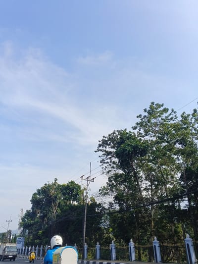 A bright blue sky with wispy clouds overhead. Green trees and foliage are on the right side of the image. Power lines are visible. A road with a white and black fence runs in the foreground. A white truck and a scooter can be seen on the road. A person wearing a blue shirt and white helmet is positioned behind a white object in the foreground.