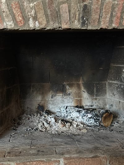 This image displays the interior of a brick fireplace, situated in the city of Sighnaghi, Georgia. The fireplace opening is topped by an arch constructed from reddish-brown and gray bricks, joined by mortar. The interior walls of the firebox are composed of dark, soot-stained bricks, predominantly black and gray, indicating extensive past use. The hearth, or floor of the firebox, is covered in a thick layer of light gray to white ash, interspersed with smaller dark charcoal fragments. A single, partially consumed wooden log lies horizontally within the ash towards the right side of the hearth. The log is largely encrusted with white ash, revealing patches of black charring. The right end of the log displays a clean, unburned cross-section, exposing its wood grain. The scene is static, devoid of active fire, smoke, or human presence.