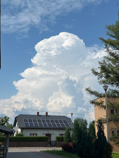 The image depicts a cloudy sky over a residential area in Borowa, Poland. The dominant subject is a large, fluffy cumulus cloud formation filling a significant portion of the frame. The sky is a clear blue with scattered white clouds.

In the foreground, the tops of trees with green needles are visible, extending into the frame from the left. Below the clouds and trees, a modern house is partially visible. The house has a dark gray tiled roof, and on the roof, a panel of solar panels is installed, reflecting the sunlight. Two red-brick chimneys protrude from the roof. The house's facade is white, with a few windows and an entrance door. A neatly trimmed green hedge runs along the front of the house.

To the right of the house, a paved walkway or driveway is visible, bordered by a wooden fence and more greenery. A lamppost with a modern design stands near the house, its light fixture extending over the property. Another lamppost is visible in the distance, also illuminating the street.

The time of day appears to be daytime, given the bright sunlight illuminating the clouds and the scene. The weather is partly cloudy with a mix of large, dramatic clouds and smaller scattered ones, suggesting a dynamic sky. The presence of solar panels indicates a focus on renewable energy. The scene conveys a sense of a peaceful, suburban environment on a sunny, but cloudy day. No people are visible in the image. There is no visible text.