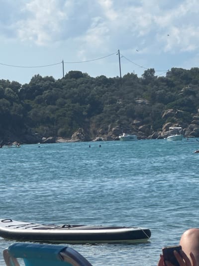 The image depicts a serene coastal scene in Ammouliani, Greece. The foreground features a portion of a paddleboard floating on calm, blue water, with a blue chair partially visible. In the mid-ground, the clear turquoise sea stretches towards a rocky shoreline covered in dense green trees. Several boats are anchored in the water, including two white motorboats. A few people are visible in the water, some appearing to be swimming or paddleboarding. In the background, a line of telephone wires stretches across the frame, supported by poles on the forested hillside. The sky is a mix of blue and white clouds.