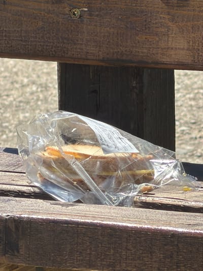 A plastic-wrapped sandwich sits on a wooden bench. The sandwich is partially visible through the clear plastic packaging, revealing layers of bread and filling. The bench appears to be weathered and outdoors, with blurred, out-of-focus greenery in the background. A label is attached to the plastic wrap, but the text is mostly illegible. The scene suggests a casual, possibly picnic-like setting. The location is Dinkata, Bulgaria.