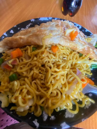This is a close-up, overhead shot of a plate of instant noodles, likely Maggi, served with an omelet on the side. The noodles are a bright yellow and appear to be mixed with chopped carrots, green peas, and thinly sliced onions. The omelet is golden brown and folded over a portion of the noodles. The plate has a black rim with a white speckled pattern. The entire dish is placed on a wooden table, with a dark-colored bottle to the left of the plate, which appears to be a bottle of soy sauce or a similar condiment. The lighting suggests it is daytime, and the overall scene depicts a casual meal being prepared or consumed. The setting is likely a restaurant or home kitchen in Ropp, Nigeria, given the provided location context. No text is visible in the image.