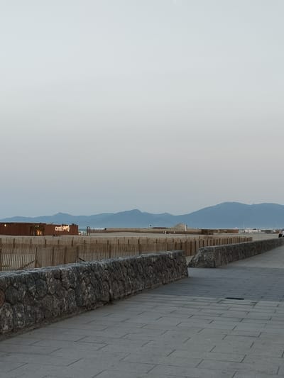 The image shows a view of the beach area in Canet-en-Roussillon, France. A paved walkway runs alongside a stone wall, which separates it from a sandy beach area. The beach is sectioned off by wooden fencing. In the background, there is a building with the illuminated text "COTE D'OPAL" visible. Beyond the beach and fencing, there are mountains in the distance under a hazy sky. The overall scene suggests a public space near the coast, possibly a promenade.