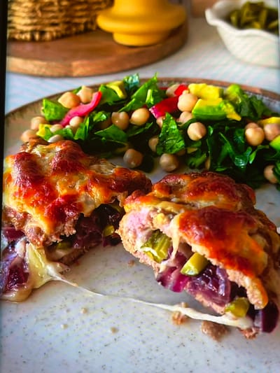 This media file shows a close-up, overhead view of a plate of food, likely a meal from a restaurant. The meal consists of two halves of a stuffed bread or pastry, with a side salad. The stuffed bread is golden brown and appears to be filled with a mixture of meat, red onion, and pickles. Melted cheese is oozing out from the sides, suggesting it was baked or broiled.
The salad on the left side of the plate contains leafy greens, chickpeas, avocado, and thinly sliced red radish or red onion. There's also a small white bowl with chopped green pickled items in the upper left corner, possibly to accompany the main dish.
The background is out of focus but suggests a table setting with a textured placemat or napkin. The overall impression is of a well-prepared and appetizing meal. The lighting is bright, indicating it's likely daytime. There are no visible people or text. The context provided suggests the location is Nijmegen, Netherlands.