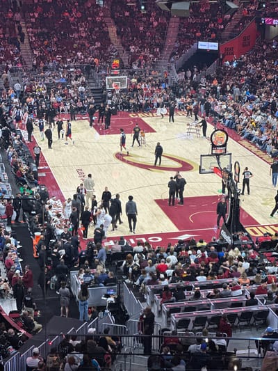 This image depicts the interior of Rocket Mortgage FieldHouse, a sports arena in Cleveland, Ohio, USA. The arena is crowded with spectators, indicating an event is either about to begin or is ongoing. The court is visible, marked with the Cleveland Cavaliers' logo and colors. Players and staff are present on the court. Scoreboards with the "T+TISSOT" logo are visible above the court. Advertising banners for brands such as Cleveland Clinic, ORedfin, Seat Geek and Michelob ULTRA are displayed around the arena. The overall scene suggests a vibrant and energetic atmosphere at a sporting event in Cleveland.
