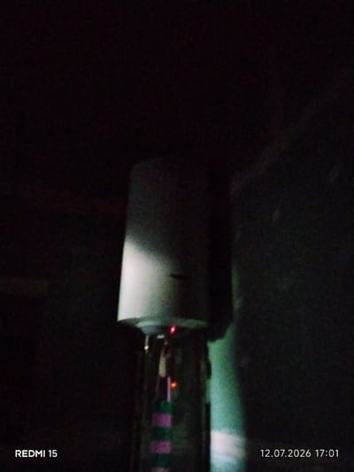 The image displays a vertical, cylindrical object, possibly a water heater or a piece of industrial equipment, set against a dark, underexposed background. A small red light is visible near the top of the object. The bottom portion of the object has horizontal stripes of pink and green. Text overlays in the bottom left corner read "REDMI 15", and in the bottom right corner, "12.07.2026 17:01". The lighting suggests an indoor or dimly lit environment. There is no clear indication of a specific location within the city of Ayiet, Kazakhstan.