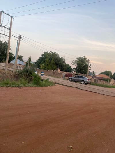 The image shows a street scene in Wauro Jabbe, Nigeria. A grey sedan is driving on a paved road. The road is lined with concrete walls and vegetation. Overhead power lines stretch across the sky. The foreground is a dirt road. The sky is overcast with some wispy clouds. A signpost is visible with directions to "Jada" and "Wuro."