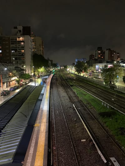 The image shows a night view of a train station in Ramos Mejía, Argentina. The perspective is from an elevated position, looking down on multiple train tracks curving into the distance. Adjacent to the tracks is a train platform with yellow tactile paving along the edge. Buildings are visible on both sides of the tracks, illuminated by streetlights and windows. The sky is dark and overcast. Cars are parked along the street running alongside the platform. The train tracks are surrounded by gravel and some vegetation.