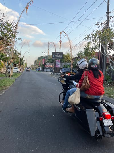 A rear view of two people on a black motorcycle on a paved road in Tabanan, Indonesia. The motorcycle is a Harley-Davidson with a license plate that reads "DK 84 JA 05-30". The passenger, in a red jacket and blue jeans, is wearing a black helmet. The driver is wearing a white helmet. Both wear helmets. The motorcycle has saddlebags and a passenger backrest. The road is lined with trees, buildings, and decorative arches strung with fabric and ornaments. A white car is parked on the left side of the road. The sky is blue with some clouds.