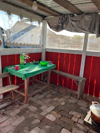 This is an outdoor covered patio area, likely in a rural setting in Покров ауылдық округі, Kazakhstan. The floor is paved with weathered bricks. A rustic wooden table covered with a green plastic tablecloth is set with a bottle of "Напитки Дюшес" soda, a pink mug, a small vase of white flowers, a pack of cigarettes, matches, a spoon, an ashtray, a small white bowl, and a green bowl. A wooden chair is pulled up to the table, and a simple wooden bench is positioned along the red wall. The back wall is painted bright red, and a corrugated metal roof is supported by wooden beams. Sheer lace curtains hang across the opening, and a dark grey drape is pulled across a portion of the roof. Sunlight casts distinct shadows on the red wall, indicating it is daytime. The overall impression is of a functional, modest outdoor living space.