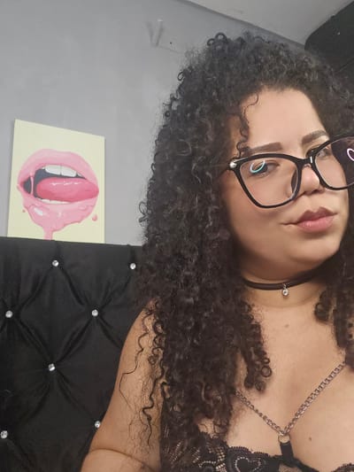 A woman with dark, voluminous curly hair is captured in a close-up portrait, lying down and looking towards the camera. She wears large black-framed glasses, reflecting a soft light source. Her eyebrows are well-defined, and her lips are slightly parted, adorned with light pink lipstick. Around her neck is a simple black choker with a small silver charm, and a silver ear piercing is visible near her ear. She is dressed in what appears to be a black lace top or bra, featuring an intricate design with silver chains draping across her chest.
The setting appears to be indoors. To her side, a section of a black upholstered surface with a diamond tufting pattern and silver stud embellishments is visible, possibly a headboard or cushion. Behind her, a plain light grey wall serves as a backdrop. On this wall, a distinctive piece of art is displayed, depicting open pink lips with a pink tongue protruding, surrounded by pink drip-like elements. The overall atmosphere suggests a casual, posed moment. There is no visible text in the image. The location context for this media file is Perímetro Urbano Cúcuta, Colombia.
