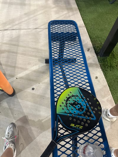 A high-angle view depicts an outdoor recreational area in McAllen, United States of America. A blue, expanded metal mesh bench extends horizontally across the frame. On the bench, a black, blue, and lime green Dunlop Blitz Attack padel racket rests, displaying its perforated face, "BLITZ ATTACK" branding, and "GRAPHITE OPEN FRAME" text. A clear plastic water bottle with a red cap is positioned near the racket. The ground consists of light grey concrete pavement. To the right of the bench, a strip of green artificial turf is visible, along with a black vertical support post. Two individuals are partially visible at the lower edges of the frame: on the left, a foot wearing a grey Nike athletic shoe with pink accents and a white sock; on the right, a foot wearing a white Nike athletic shoe. Shadows are cast on the concrete, indicating an external light source.