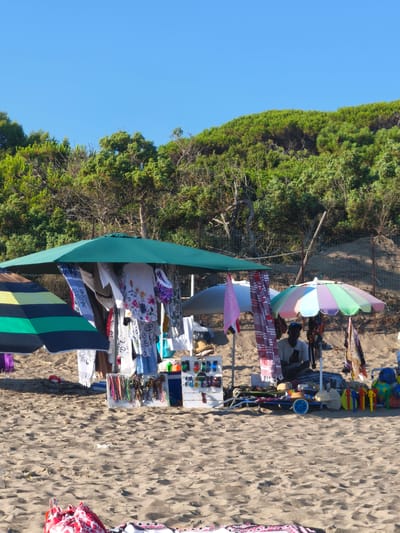 In Castiglione della Pescaia, Italy, a beach vendor operates a stall on a sandy beach, selling various beachwear and accessories. The stall is set up under a large green umbrella, with clothing items like shirts and scarves displayed on a rack. Smaller items such as sunglasses, hats, and necklaces are arranged on tables and shelves. A man, presumably the vendor, is seated behind the stall, looking at his phone. To the left of the main stall, another umbrella with colorful stripes provides shade, and a separate display features more clothing. The background shows a dense green hillside covered in trees and shrubbery, with a clear blue sky above. The sandy ground is partially covered with scattered items and footprints, indicating the presence of beachgoers.
