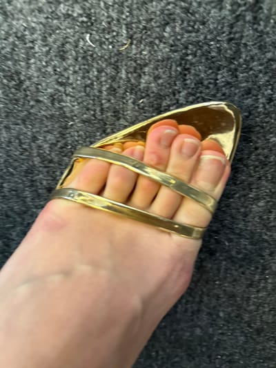 A single, fair-skinned human left foot is depicted wearing a gold-colored, open-toed sandal. The scene is located in Arcueil, France. The sandal features a metallic finish with two parallel, reflective straps securing the forefoot and toes. The toes display natural toenails with unpainted, visible white tips. Minor dermal irregularities, consistent with calluses, are present on the superior surface of the second and third toes. The foot and sandal rest on a dark grey, densely textured carpet. Sparse, light-colored fibrous debris is visible on the carpet in the background.
