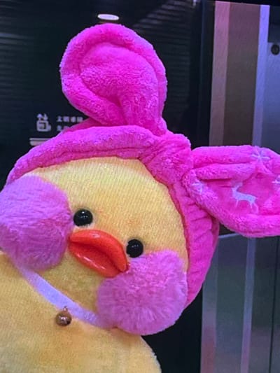 This media file is a close-up photograph of a stuffed toy chick. The chick is primarily yellow, with a pink hood that resembles bunny ears. The hood has small white star-shaped patterns. The chick has two black eyes and an orange beak. It also has pink fluffy cheeks and a pink fuzzy chin. Around its neck, it wears a thin white ribbon with a small golden bell attached. The background is dark and out of focus, but appears to be a surface with horizontal lines. There are some faint vertical lines and some Chinese characters visible in the lower left portion of the background. The lighting is artificial and casts a slight purplish hue. The image focuses solely on the toy, giving no indication of the time of day, weather, or a specific location beyond what can be inferred from the background text. No discernible activity or event is taking place.