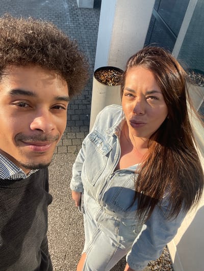 The image displays a selfie taken outdoors in Montigny-le-Bretonneux, France, featuring two individuals under direct sunlight.
On the left, a male subject with dark, curly hair and a short beard is visible from the chest up, looking directly at the camera with a smile. He wears a light-colored, possibly striped, collared shirt under a dark, crewneck sweater.
To his right, a female subject is visible, looking at the camera with a slight puckered expression. She has long, dark brown hair and is wearing a light blue denim jumpsuit with a visible front zipper. Her skin tone appears olive, and her eyes are dark.
The background consists of a paved surface, featuring both dark, textured ground and grey interlocking paver stones. Behind the subjects, two cylindrical, light grey receptacles are present; the one closer to the center is filled with numerous cigarette butts. A modern building with reflective glass panels and light-colored structural elements is partially visible in the upper right corner. Strong sunlight casts distinct shadows and highlights on the subjects and environment.