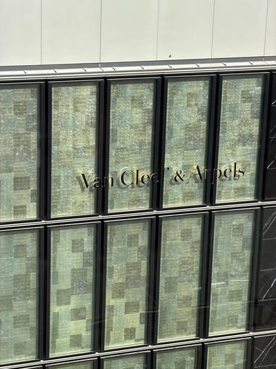 This is a high-angle, medium shot of the exterior of a Van Cleef & Arpels store in the city of Chuo, Japan. The building features a modern facade with large, vertically oriented windows. The windows are made of frosted glass with a subtle, repeating pattern of lighter and darker squares, creating a mosaic-like effect. The store's name, "Van Cleef & Arpels," is emblazoned in elegant, metallic lettering across the middle section of the facade, reflecting the sunlight. Above the windows, a section of the building is clad in white panels. To the far right, a vertical panel of black marble with white veining is visible, providing a contrasting texture. The scene is captured on a bright, sunny day, with strong shadows cast by the building's architectural elements.