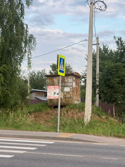 In Soloslovo, Russia, a rural road is depicted with a pedestrian crossing clearly marked by white stripes on asphalt. To the right of the road, a grey concrete pole stands tall, supporting numerous black electrical wires that loop and tangle at its top. Adjacent to this pole, another similar pole is visible, slightly shorter and further back. Beside the road and to the left of the poles, a rusty, rectangular metal shed or container sits on a grassy, elevated mound. A signpost with a blue and yellow pedestrian crossing symbol is positioned in front of the shed. Two additional signs are attached to the shed's front; one is red and white with Russian text and an image, and the other is green and white with Russian text. Beyond the shed and poles, lush green trees and foliage fill the background, with a strip of darker greenery and a brown fence further in the distance. The sky above is partly cloudy, with a mix of blue sky and white clouds.